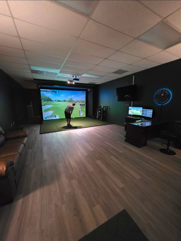 Indoor golf simulator room with large projection screen displaying a golf course, player in swing position, computer stations, and blue wall clock