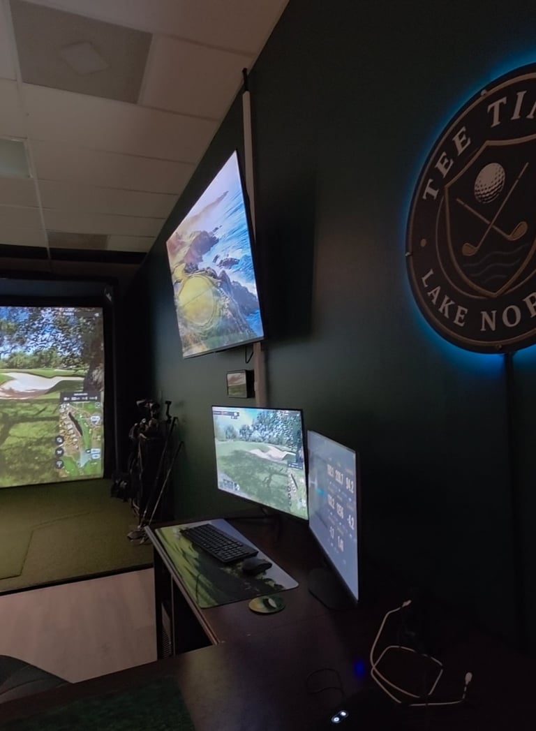 Indoor golf simulator setup with multiple monitors displaying golf course views, keyboard, and golf-themed neon sign on dark wall