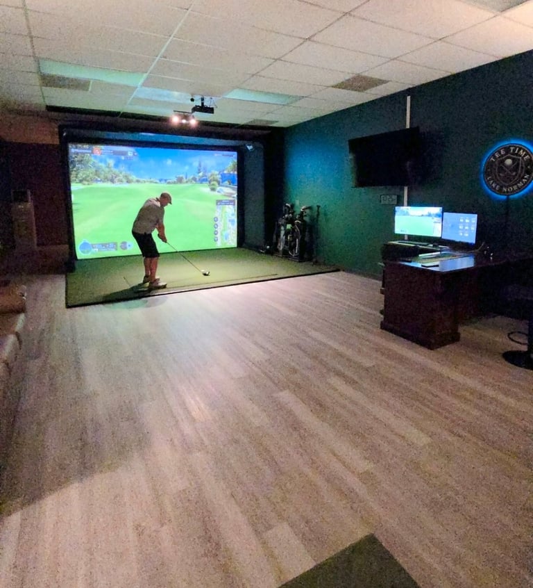 Indoor golf simulator bay with person swinging at ball while large projection screen displays golf course