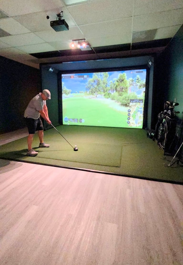 Man using golf club in indoor simulator bay with large screen displaying golf course, artificial turf, and equipment on side